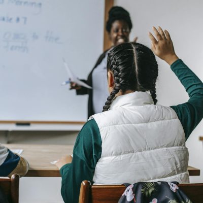 Smiling African American female teacher standing near whiteboard and looking at schoolgirl raising hand